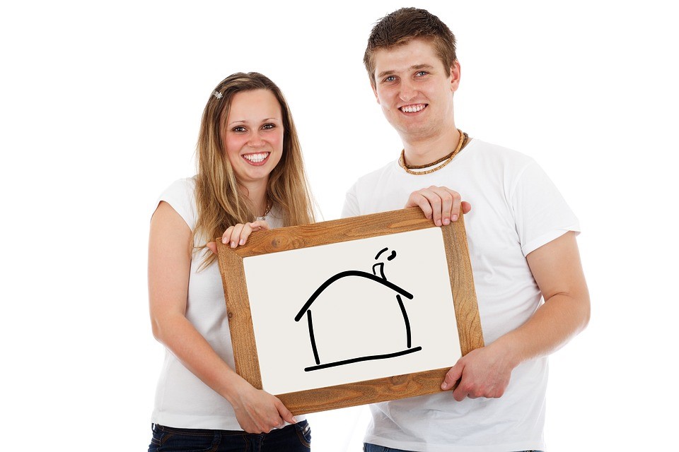 blonde man and woman wearing white t-shirts holding white board with black line drawing of a house in a white room