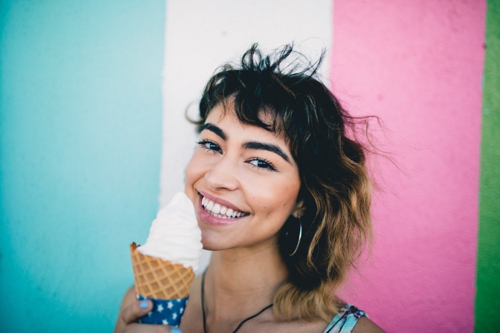 brunette woman smiling holding vanilla ice cream cone standing in front of blue white pink and green wall