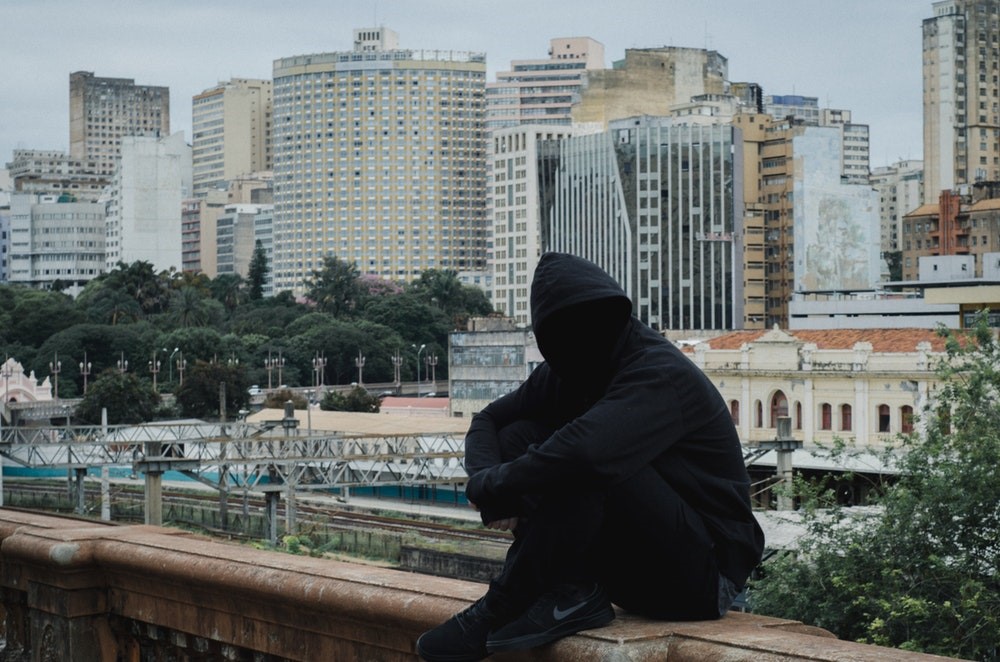 man in all black with a hoodie obscuring his face sitting on concrete in front of a city skyline