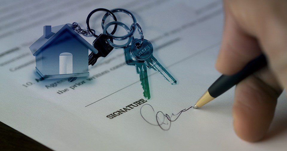 person signing a contract under the word signature with a set of keys attached to a house keychain on top of the form