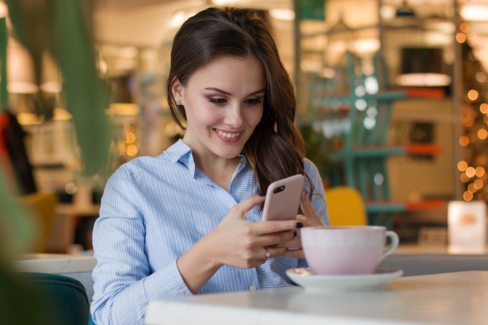 smiling brunette woman in blue dress shirt holding a phone while sitting in a café with a mug on the table in front of her