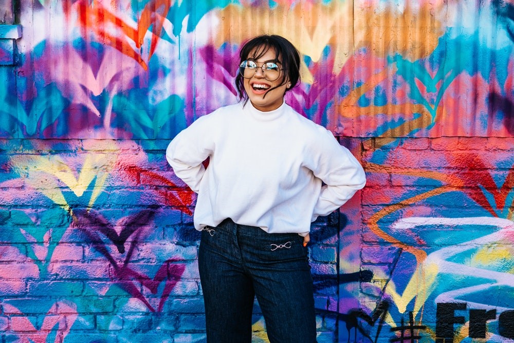 smiling woman in white mock turtle neck jeans and glasses standing in front of colorful graffiti