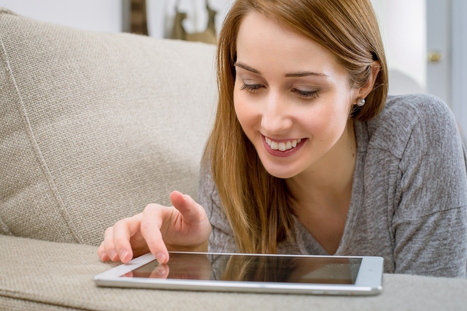 smiling woman with blonde hair using tablet with right hand resting on arm of beige fabric couch