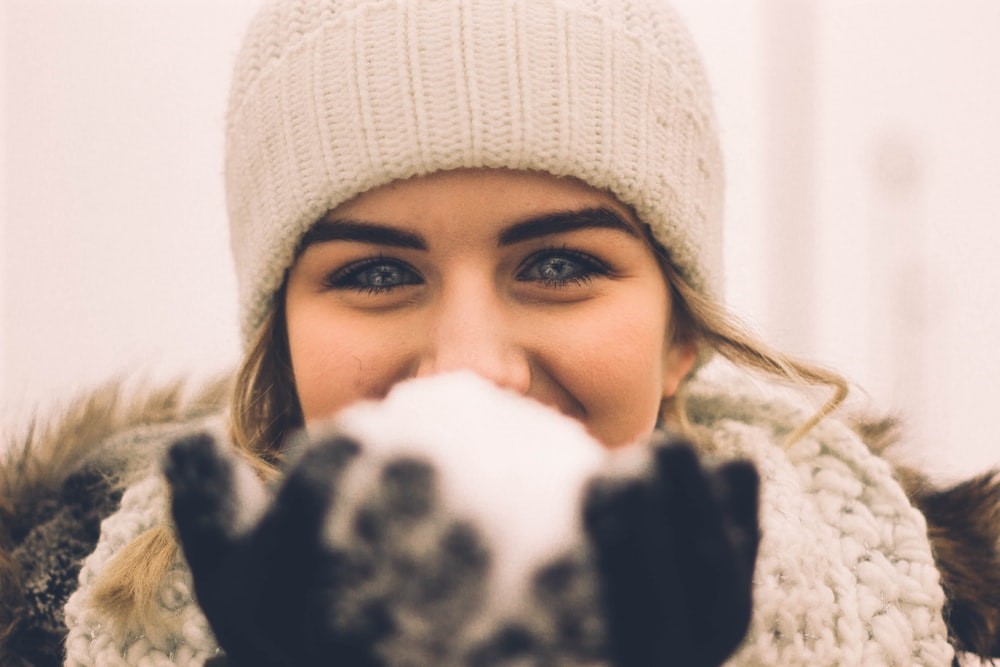 woman in knitted cream hat and scarf holding snowball in front of face