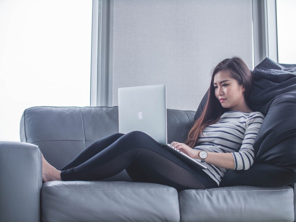woman in leggings and striped shirt sitting on sofa with laptop on her lap