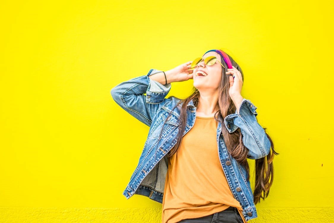 woman in orange shirt and blue jean jacket touching sunglasses looking up in front of yellow wall