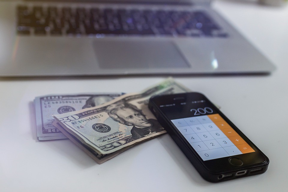 iPhone open to calculator app on a stack of American $10 and $20 bills next to a laptop