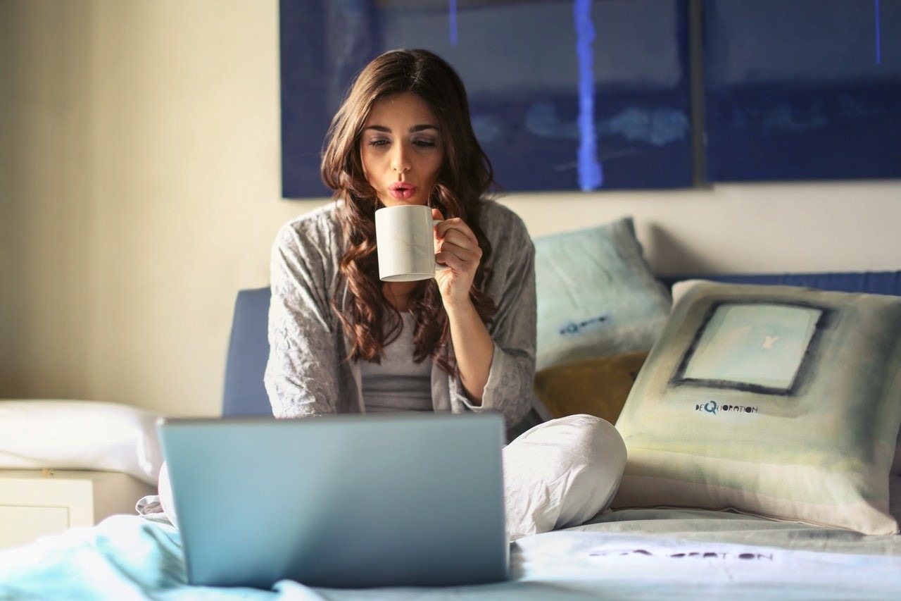 woman sitting on bed in front of open laptop holding white mug in left hand